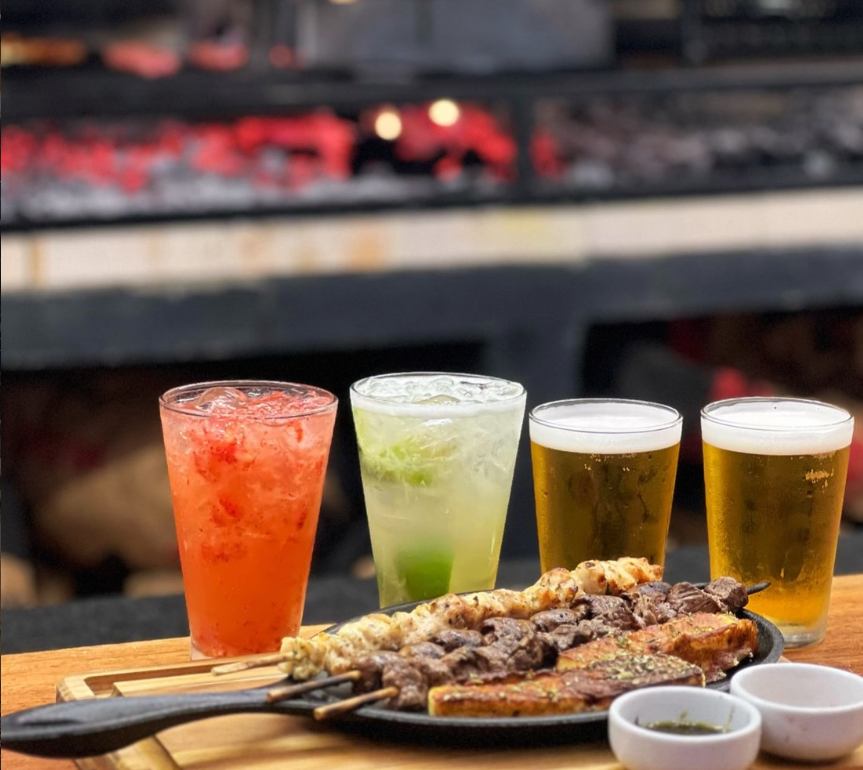 Drinks de morango e limão ao lado de dois chopes com espetinhos ao fundo da churrasqueira